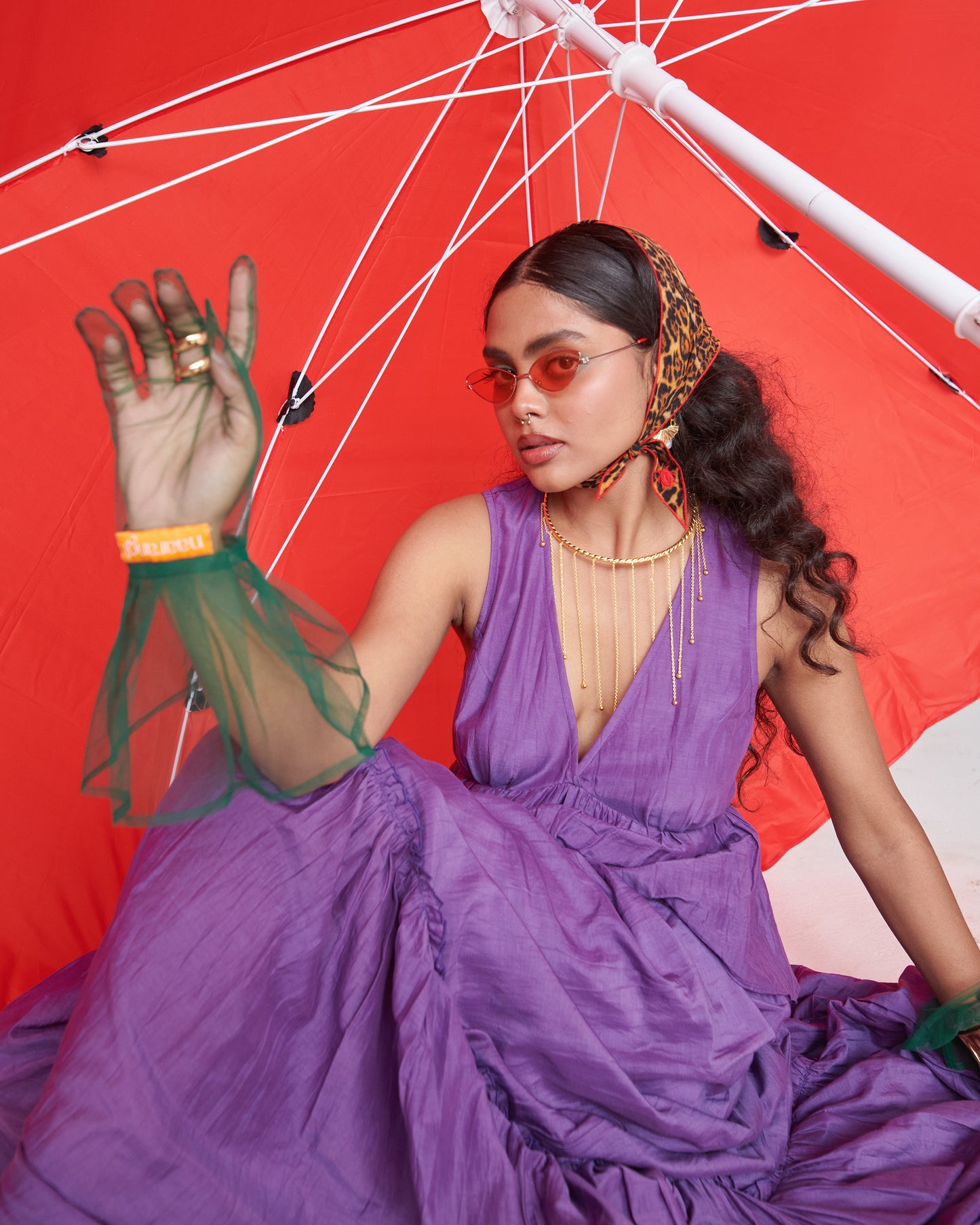 Model in KalaKhatta Crush Dress, purple layered maxi, styled with jewelry, scarf and red umbrella backdrop for fashion shoot.