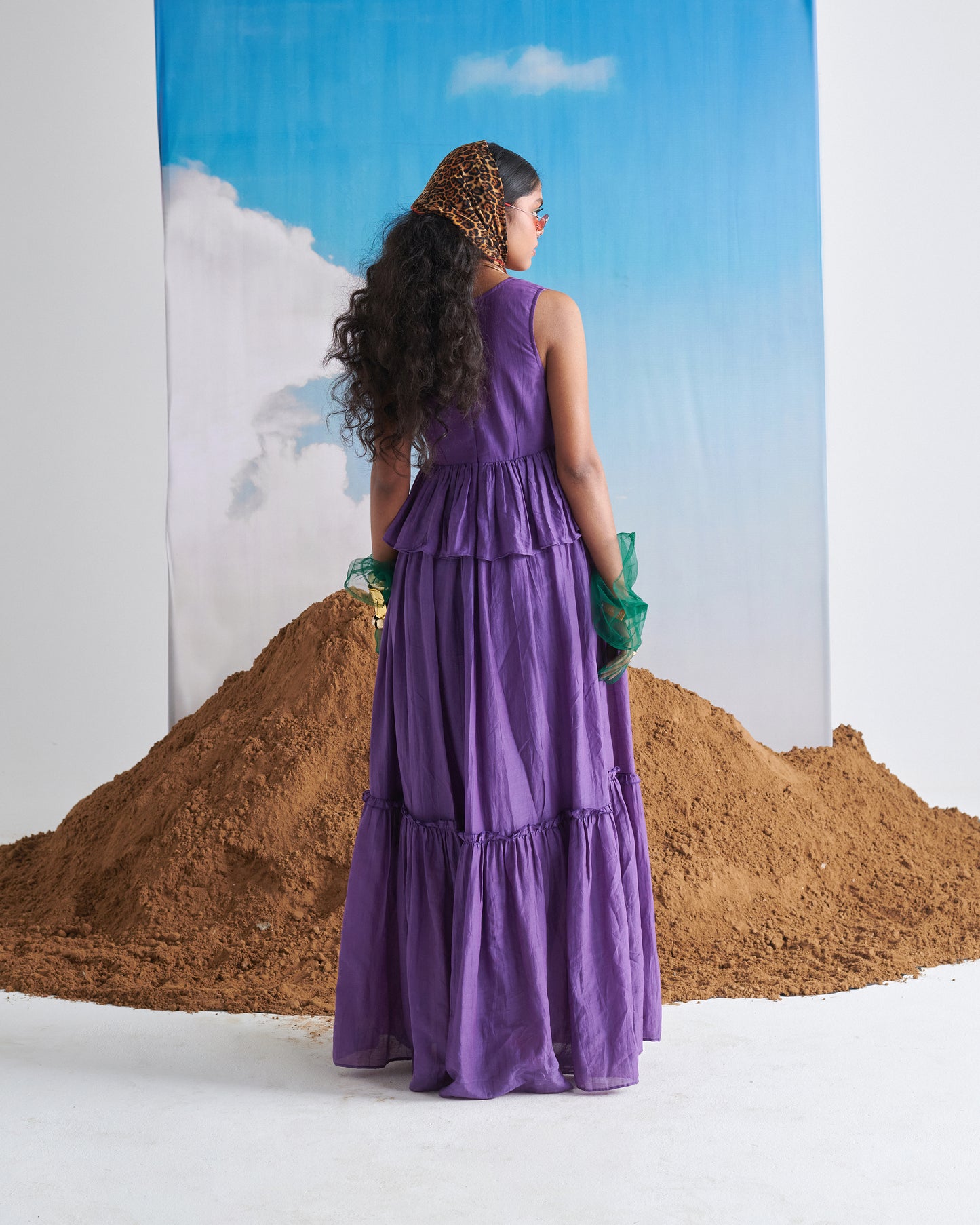 Back view of model wearing Kalakhatta purple maxi dress with green gloves, standing in front of sand mound and sky backdrop.