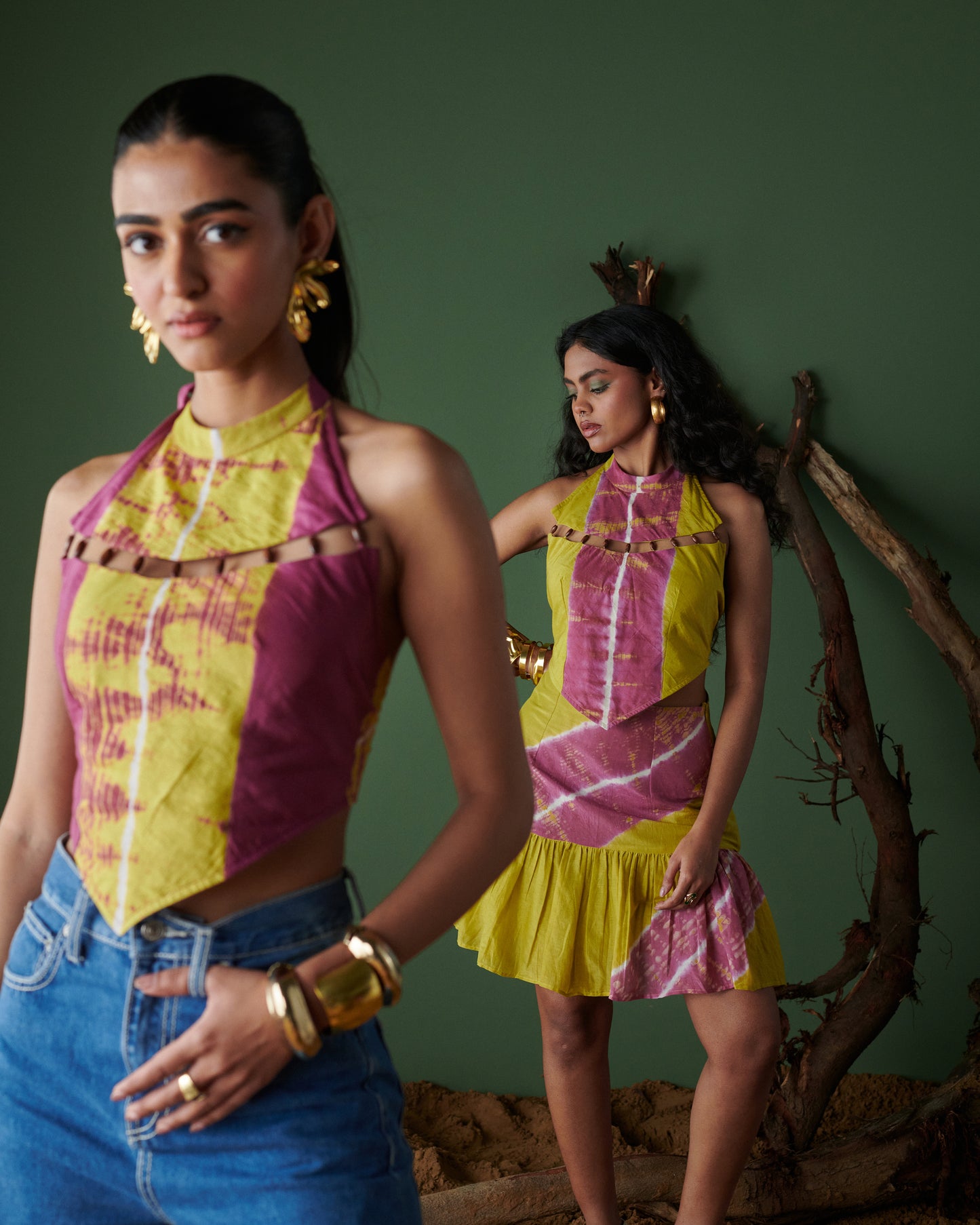 Two models in mustard yellow and magenta tie-dye halter-neck outfits, one paired with blue denim jeans and the other with a matching skirt, styled for a contemporary fashion photoshoot with a green background and rustic wooden props.