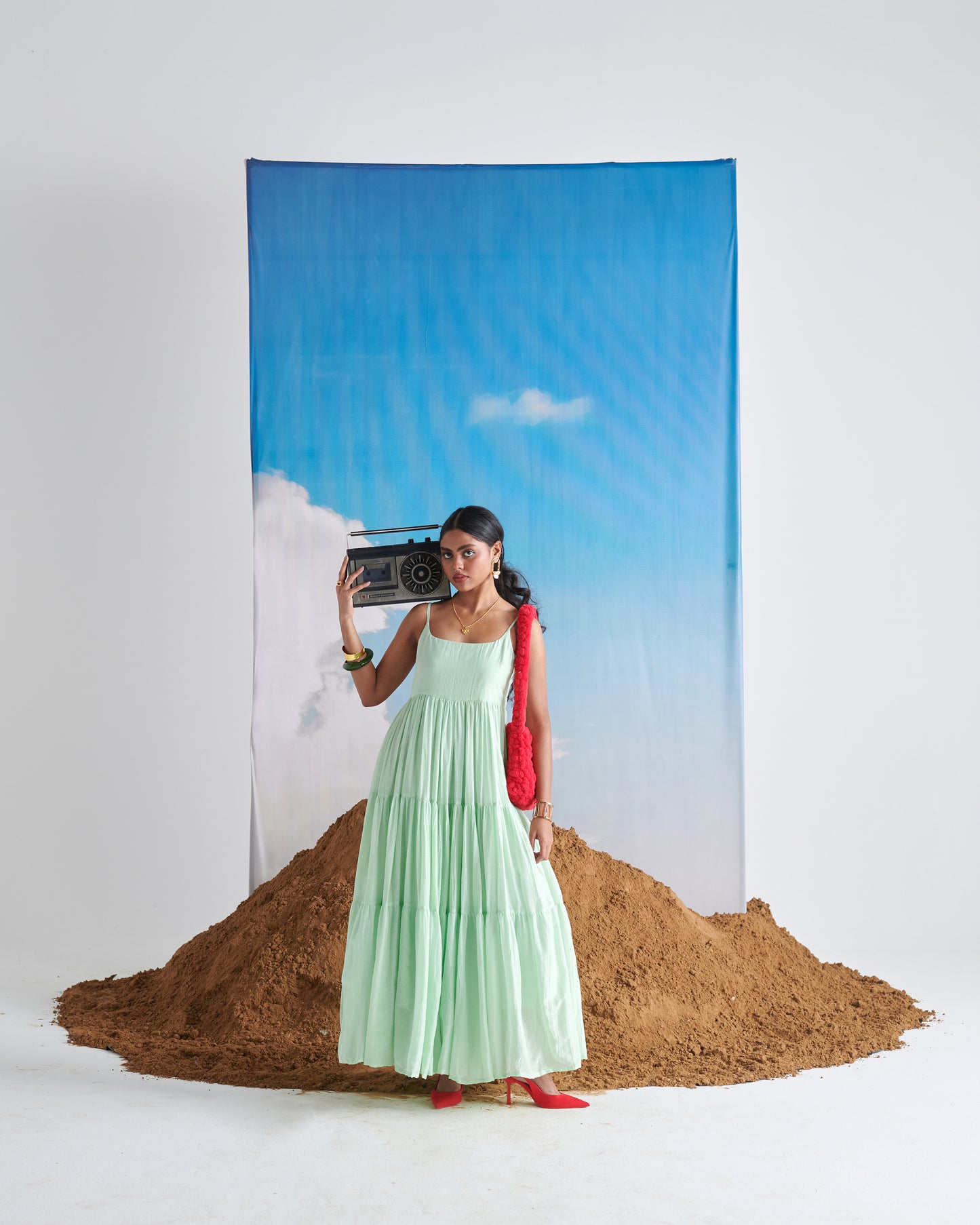 Woman in Minty Mehfil green maxi dress holding a boombox against sky backdrop with earthy mound.