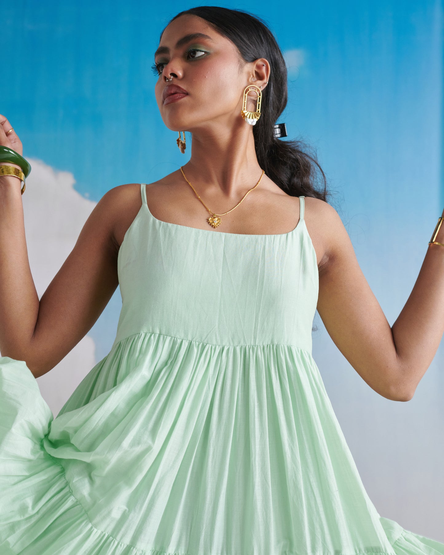 Close-up of woman in Minty Mehfil green maxi dress with bold gold jewelry and blue sky backdrop.