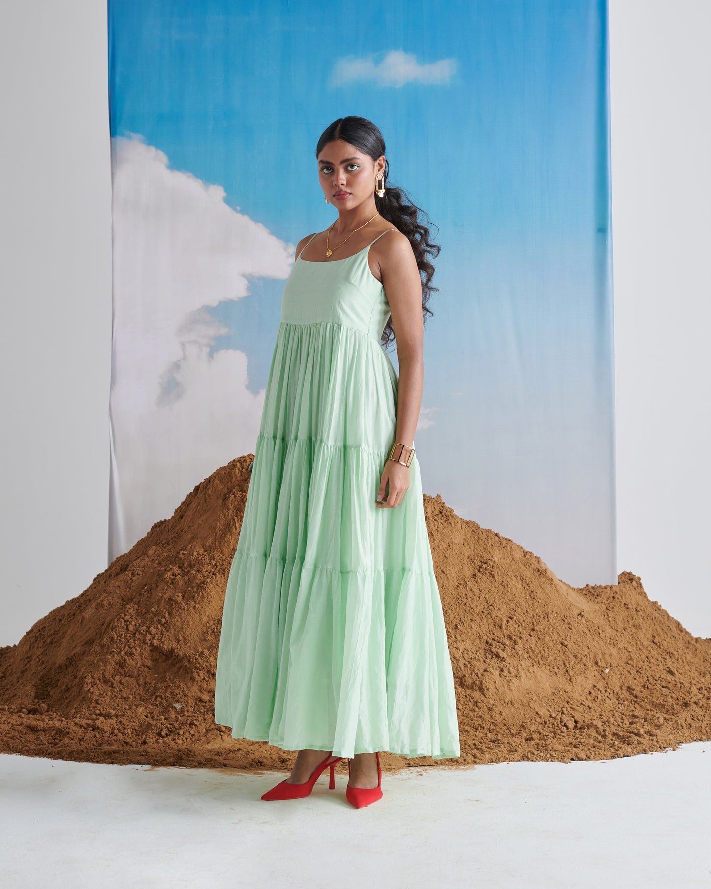 Woman in Minty Mehfil green maxi dress posing in front of sand mound with blue sky backdrop.