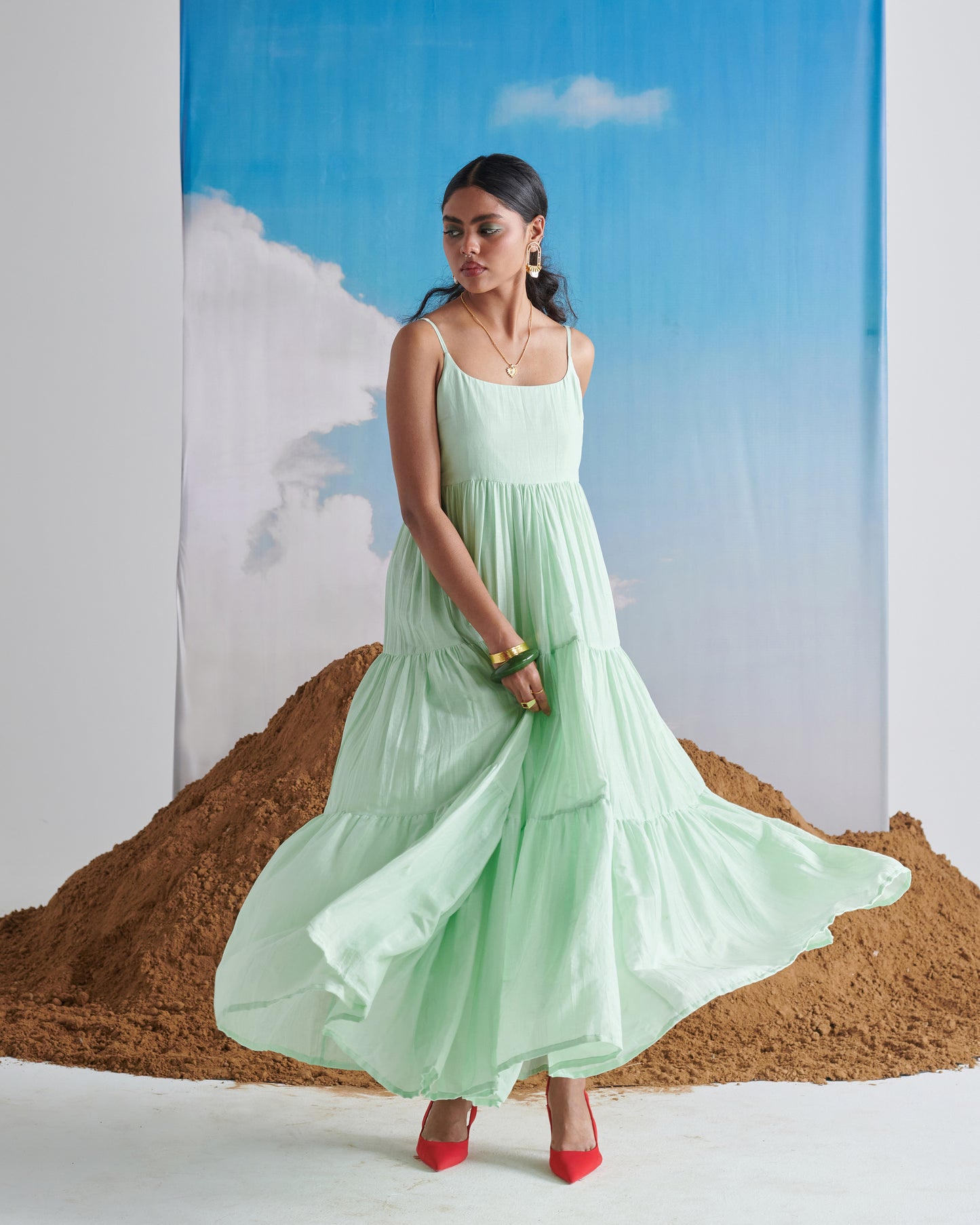 Woman twirling in Minty Mehfil green maxi dress with red heels against sand mound and sky backdrop.