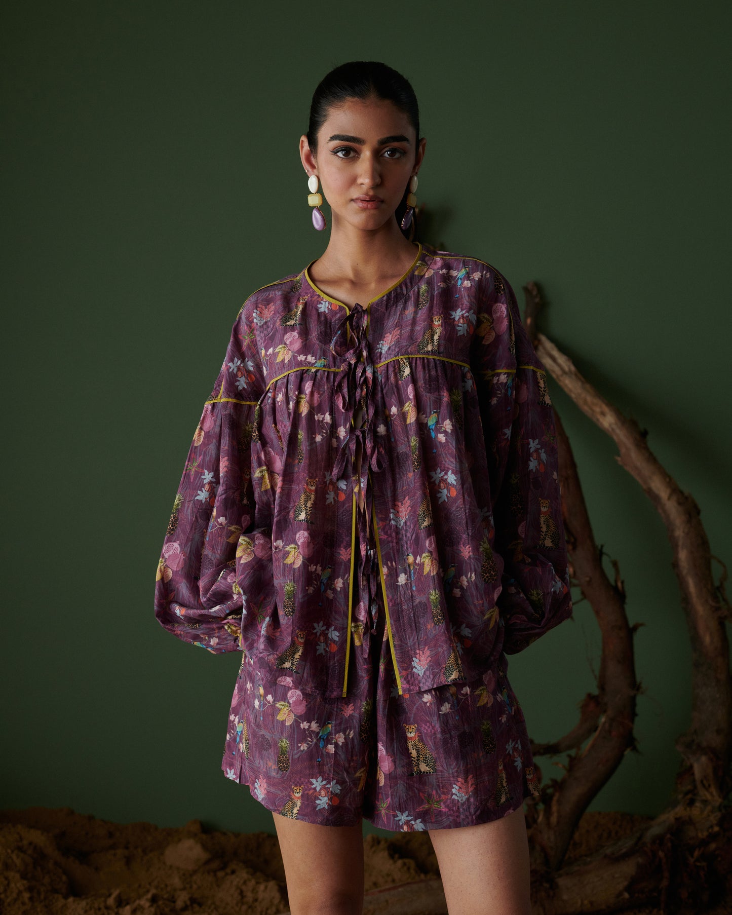 Model front view wearing Tropical Ras purple floral shorts co-ord set with long sleeves, green backdrop and earthy decor.