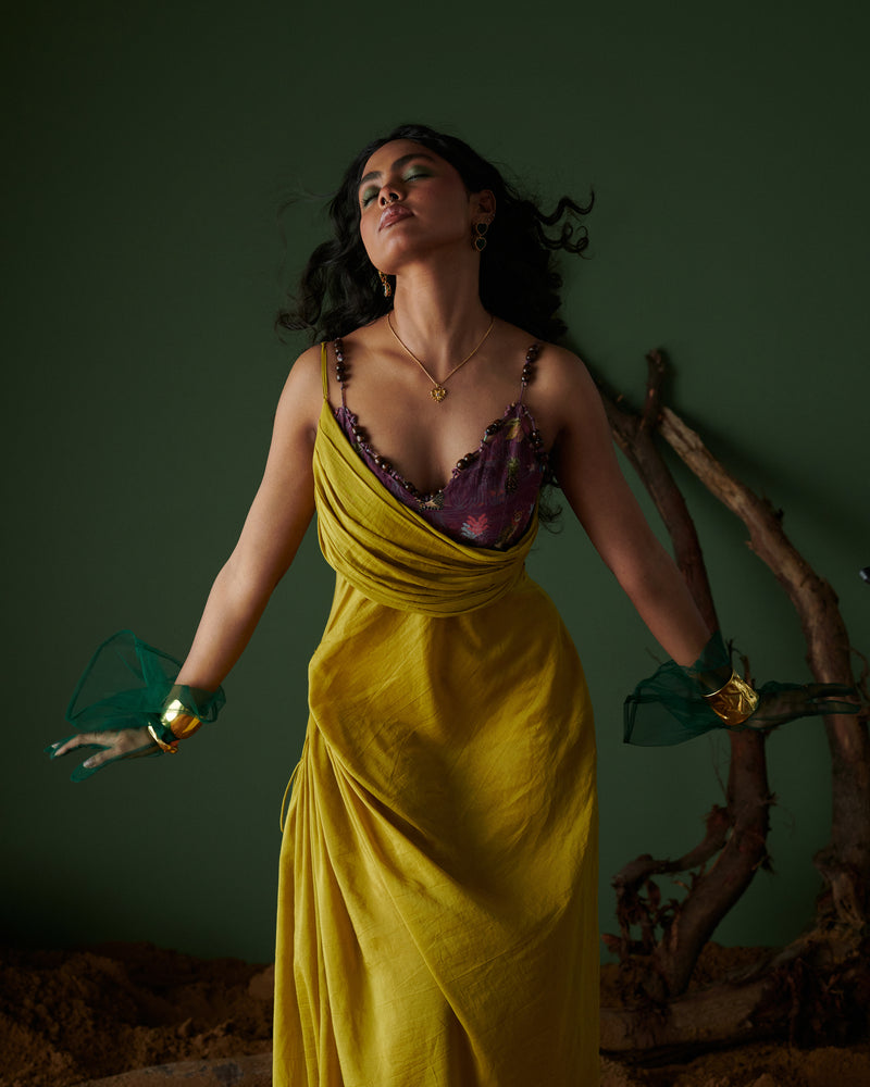 Model wearing Naarangi Sunheri Zest yellow draped dress with purple lace neckline and green mesh gloves on earthy backdrop.