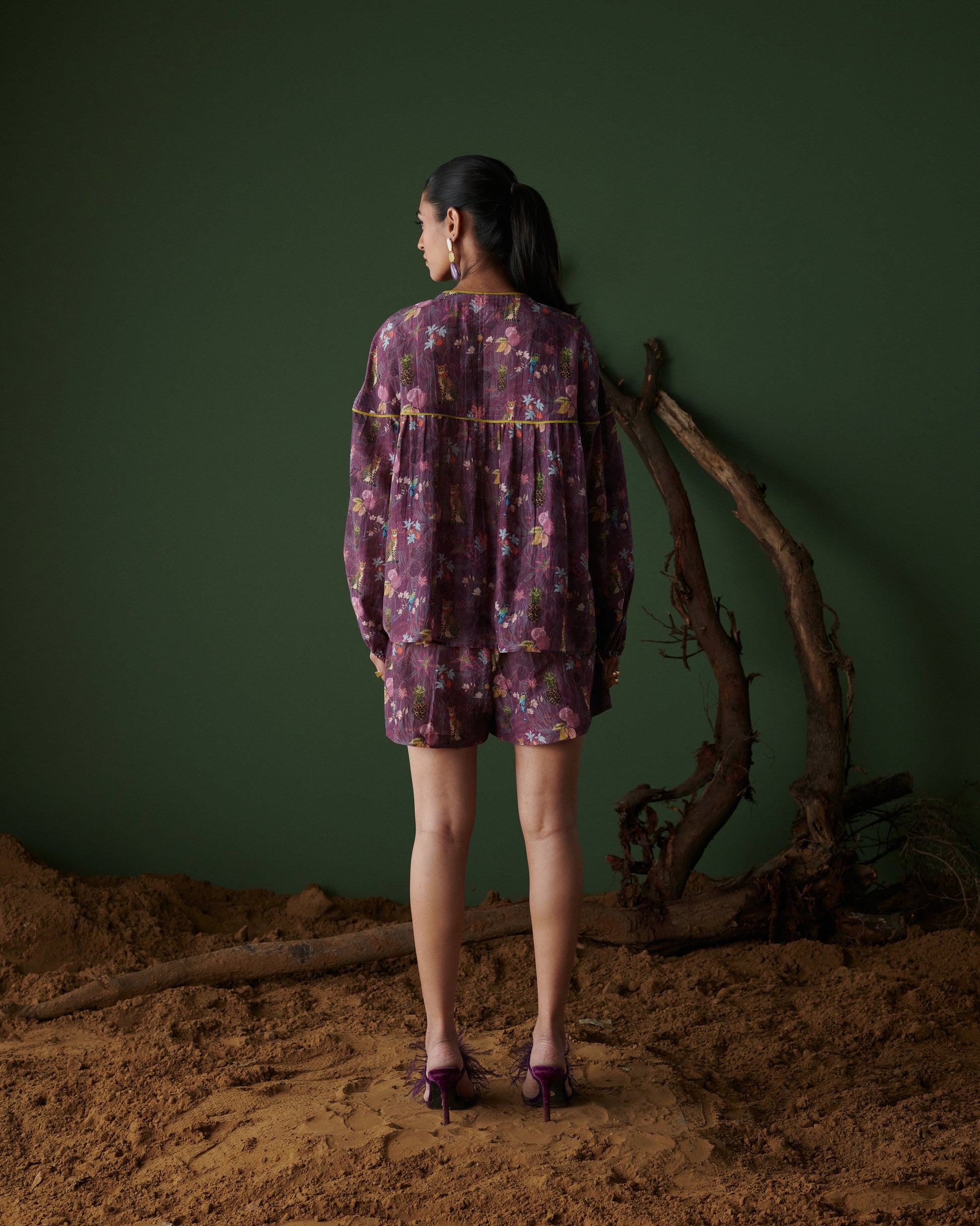 Model back view wearing Tropical Ras purple floral shorts co-ord set with long sleeves, green backdrop and earthy decor.