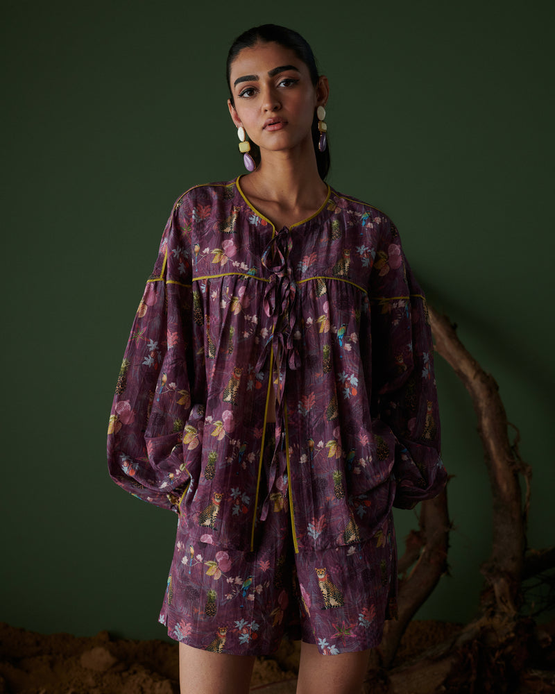 Close-up of model wearing Tropical Ras purple floral shorts co-ord set with long sleeves, green backdrop and wooden branch decor.