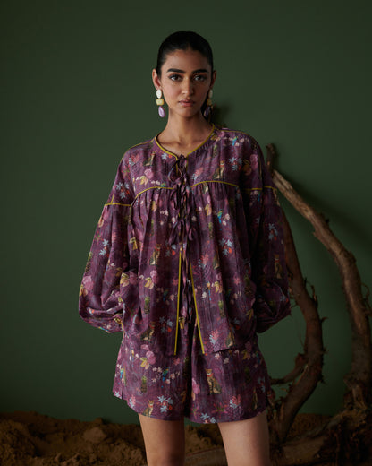 Model front view wearing Tropical Ras purple floral shorts co-ord set with long sleeves, green backdrop and earthy decor.