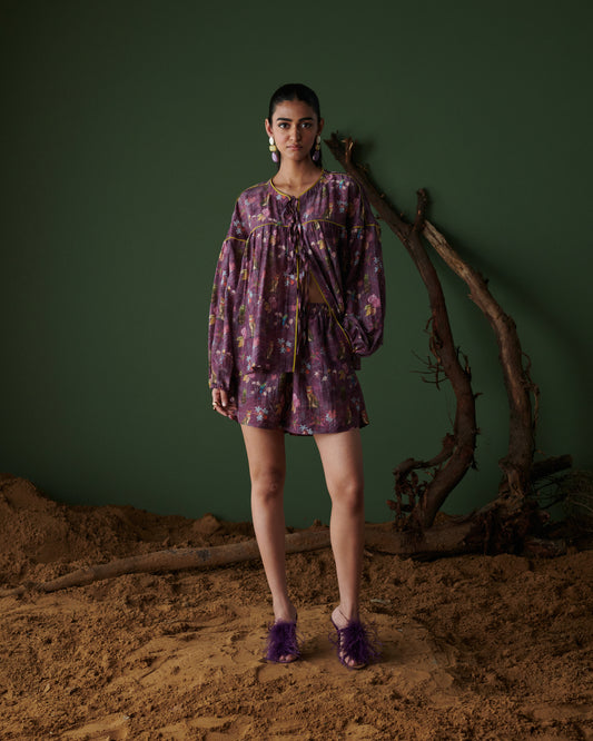Model wearing Tropical Ras purple floral shorts co-ord set with long sleeves, standing on sand with green backdrop and tree branches.