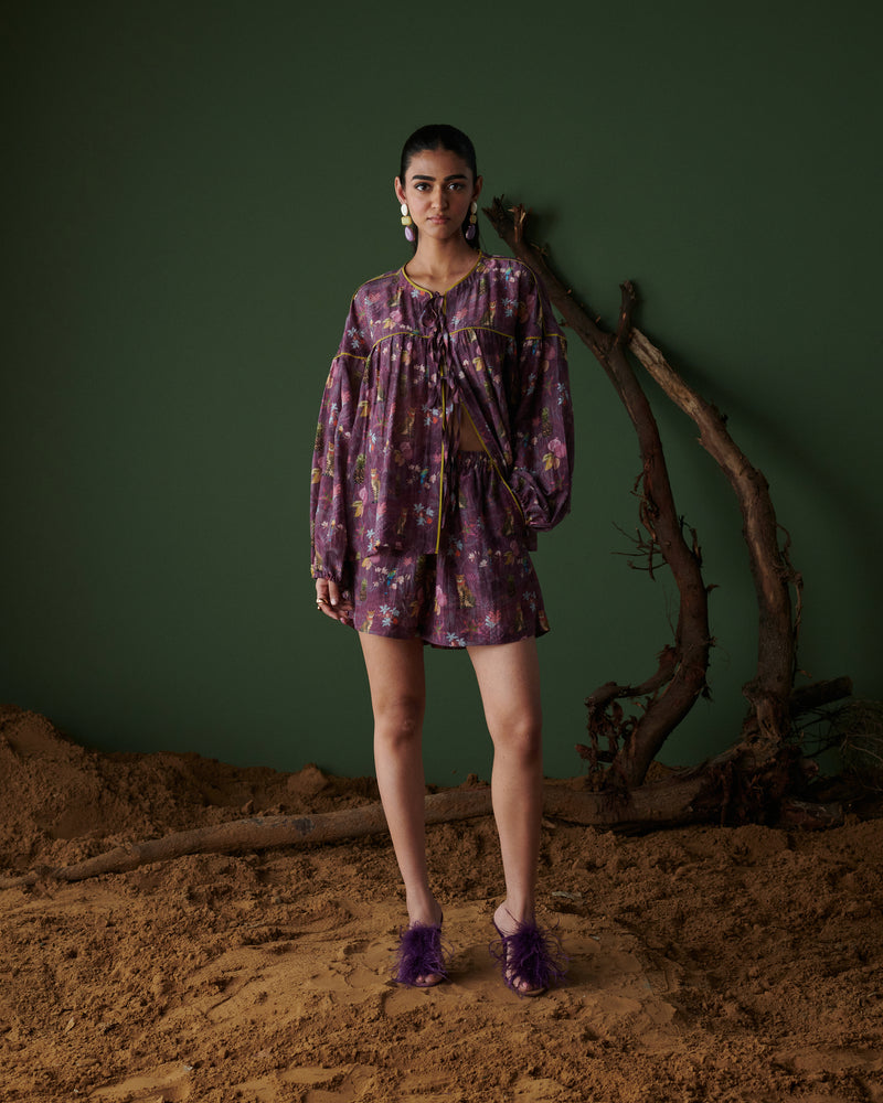 Model wearing Tropical Ras purple floral shorts co-ord set with long sleeves, standing on sand with green backdrop and tree branches.